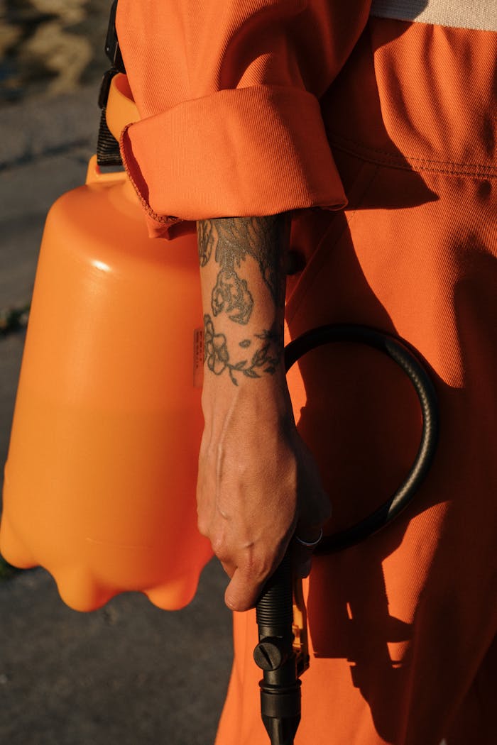 Contact Person in orange coveralls holding a plastic sprayer, focus on tattooed arm.